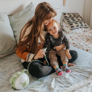 Eine Mutter sitzt mit ihrer lächelnden Tochter auf einem Bett und hält einen Nosiboo Pro elektrischen Baby Nasensauger