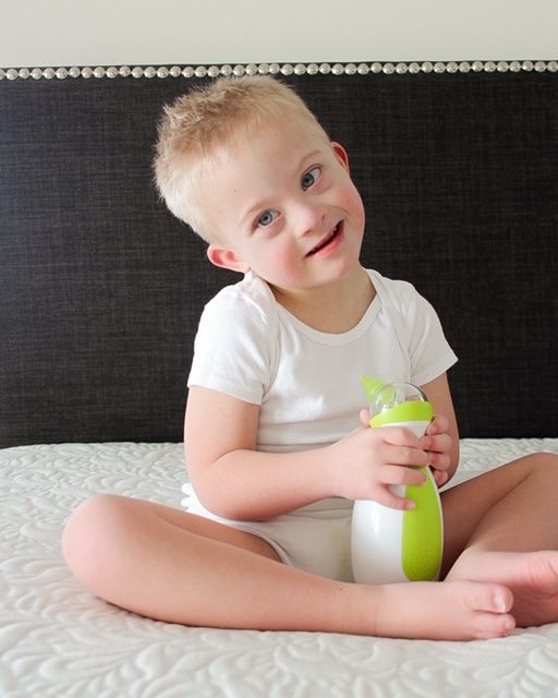 Uroczy mały chłopiec siedzi na łóżku, trzyma w dłoniach przenośny aspirator do nosa Nosiboo Go