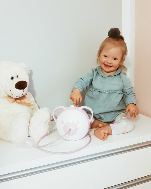 Ein lächelndes kleines Mädchen mit einem rosa Nosiboo Pro elektrischen Baby Nasensauger und einem Plüsch-Teddybär