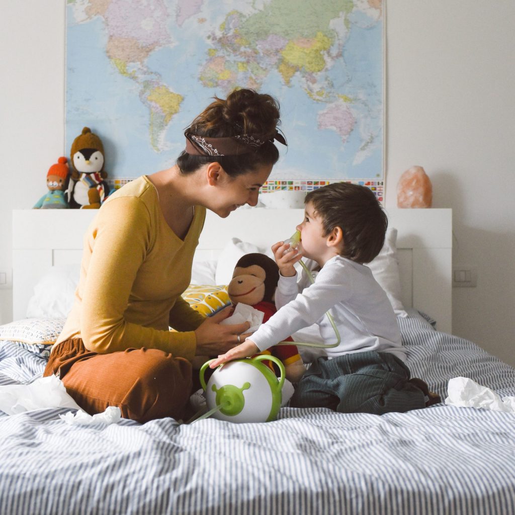 Eine Mutter sitzt mit ihrem Sohn auf einem Bett und saugt seine Nase mit einem elektrischen Nasensauger Nosiboo Pro ab