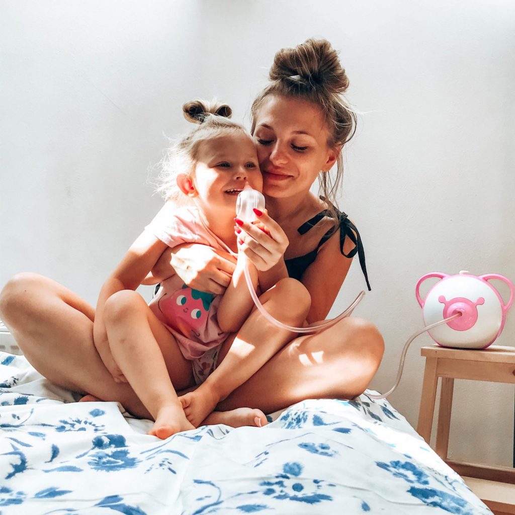 Une mère souriante et sa petite fille sont assises sur le lit et utilisent le mouche-bébé électrique Nosiboo Pro