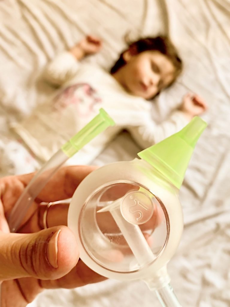 Dziecko śpi spokojnie w łóżku, nad nim ręka trzyma ustny aspirator do nosa Nosiboo Eco