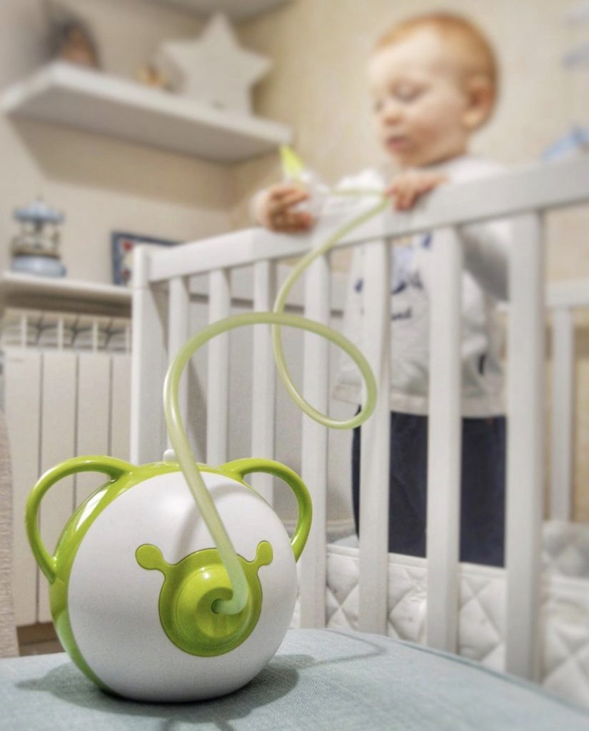 Ein grüner elektrischer Nasensauger Nosiboo Pro und im Hintergrund steht ein kleiner Junge auf einem Bett