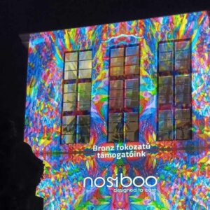 A Nosiboo logó színes fényekkel egy épület falán, ami a pécsi Zsolnay Fényfesztivál bronz fokozatú támogatásáról tájékoztat.