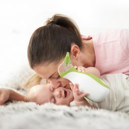 Una madre bacia suo bambino che tiene l'aspiratore nasale portabile Nosiboo Go in mano sdraiato sul letto