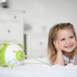 Mosolygós kislány hasalva könyököl a gyerekszobai ágyon, mellette zöld Nosiboo Pro elektromos orrszívó